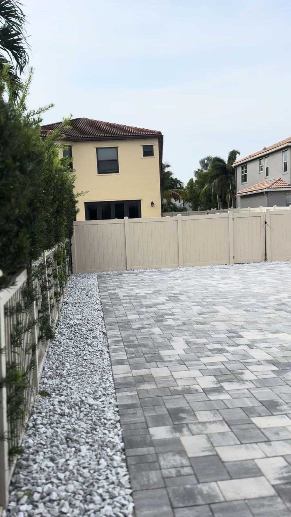 Paved outdoor space with gravel borders and a beige fence, featuring a house in the background.