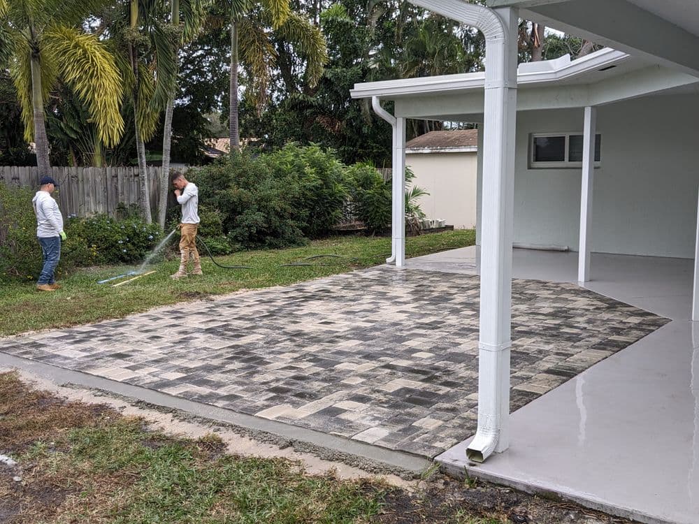 Workers cleaning a paved area in a backyard with greenery and a house in the background.