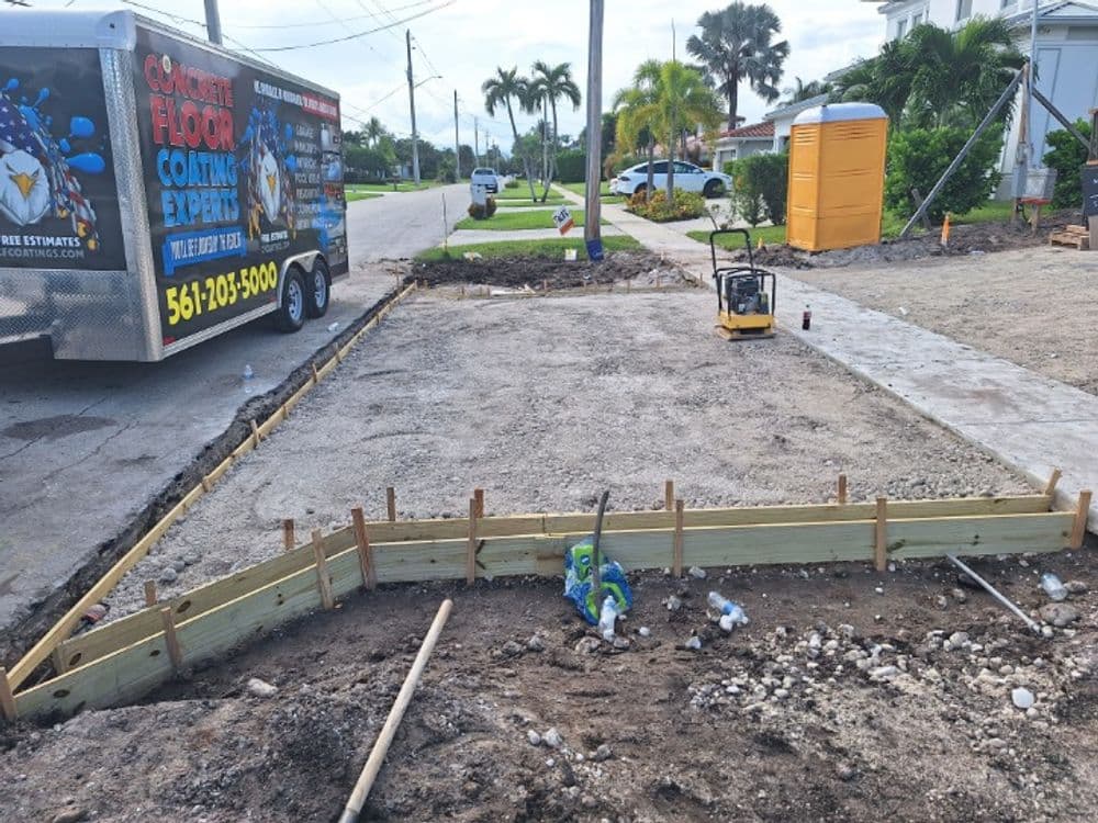 Construction site with wooden forms for concrete, paving equipment, and a trailer nearby.