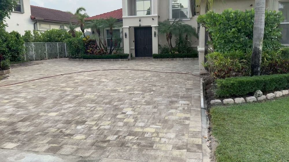 Paver driveway with landscaped gardens and a modern home entrance, featuring tropical plants.