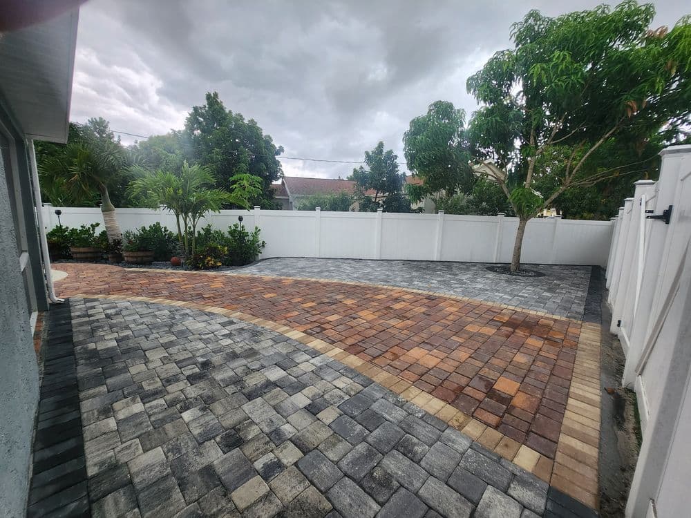 Patio with paver stones in a landscaped backyard with trees and a white fence.