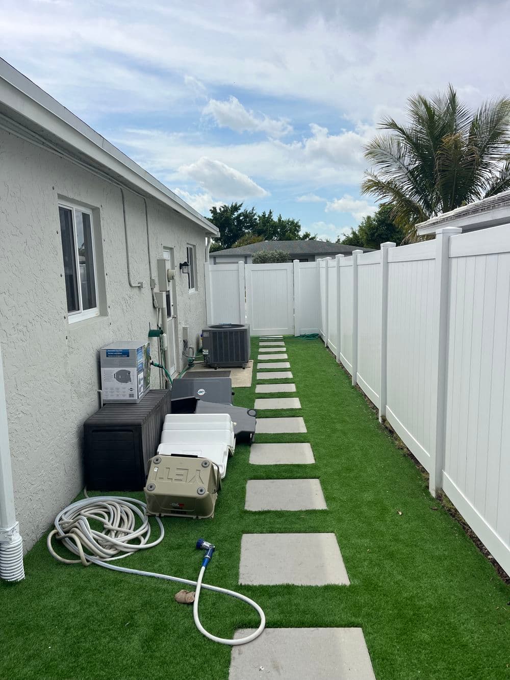 Side yard walkway with artificial grass, storage containers, and white vinyl fencing.