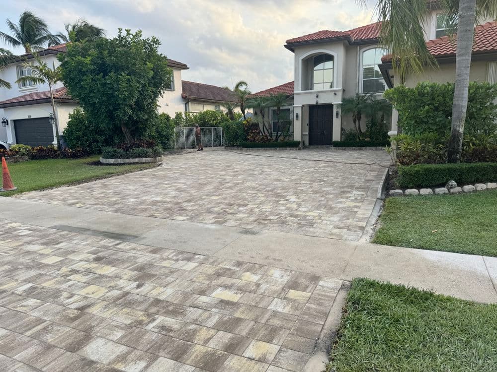 Spacious driveway with patterned pavers and tropical landscaping beside a two-story home.