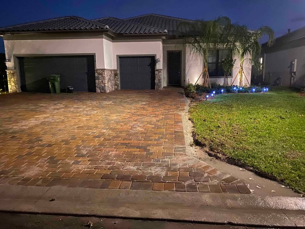Modern house exterior at night with paved driveway, palm trees, and blue landscape lighting.