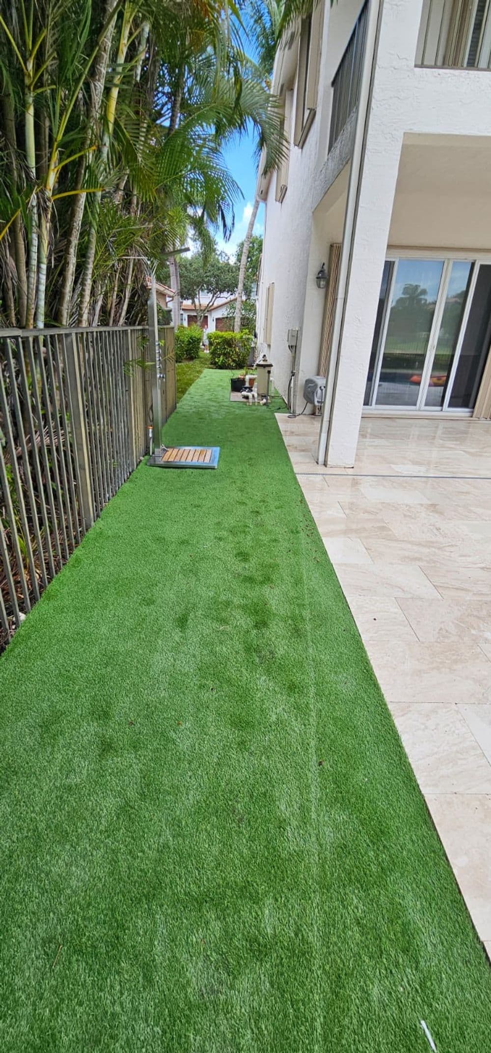 Artificial turf pathway beside a modern home with tropical landscaping and sliding glass doors.