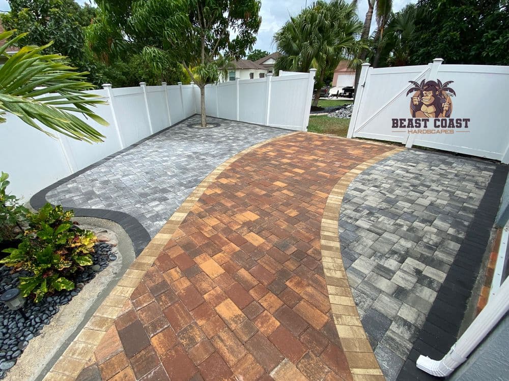 Modern stone paver driveway design with palm trees and a white fence in residential landscape.