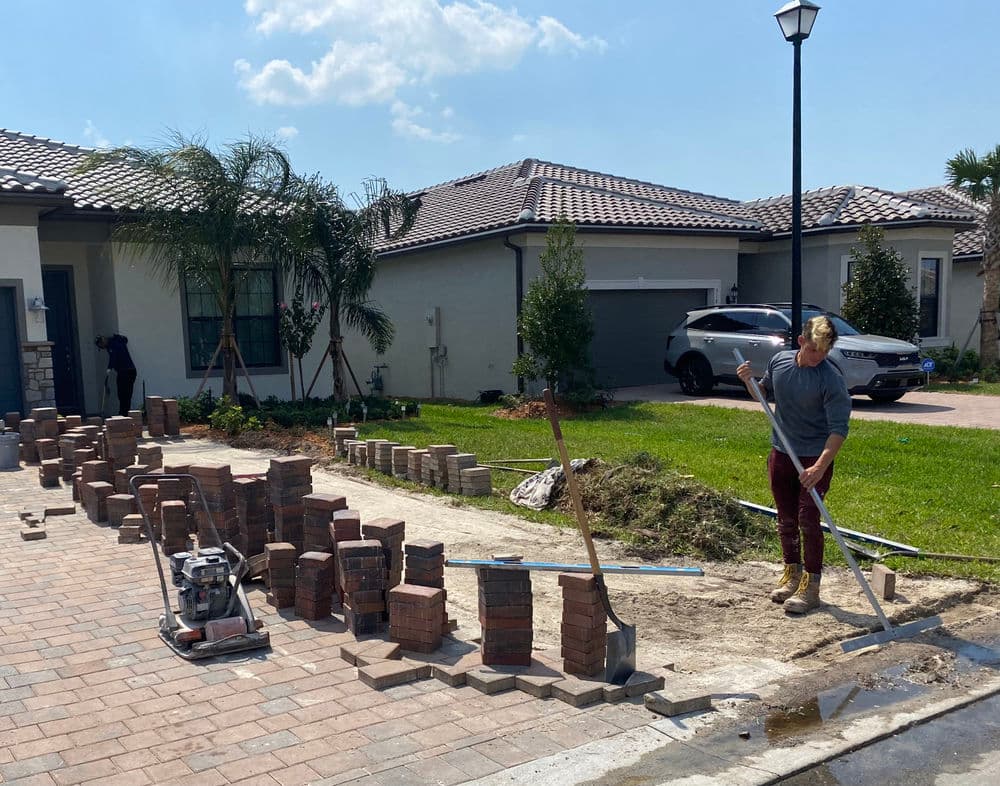 Construction worker laying bricks in a residential area with palm trees and parked cars.