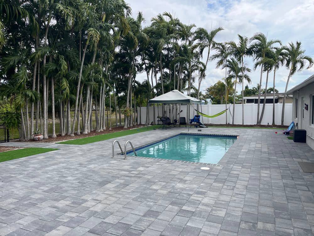 Backyard pool area with pavers, surrounded by palm trees and a shaded seating area.