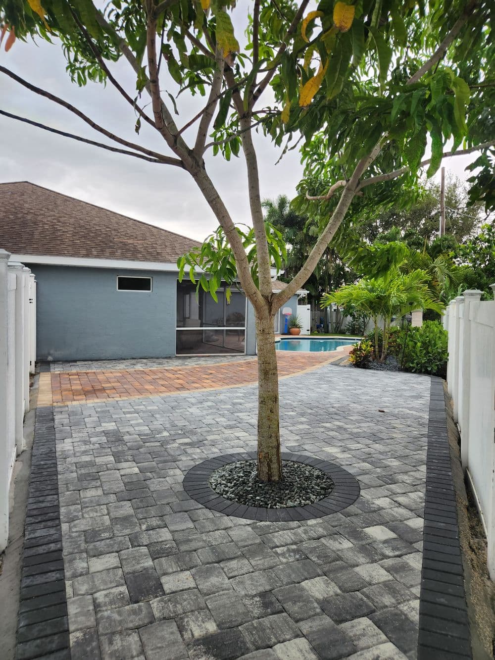 Paver stone walkway leads to backyard pool, framed by lush greenery and a central tree.
