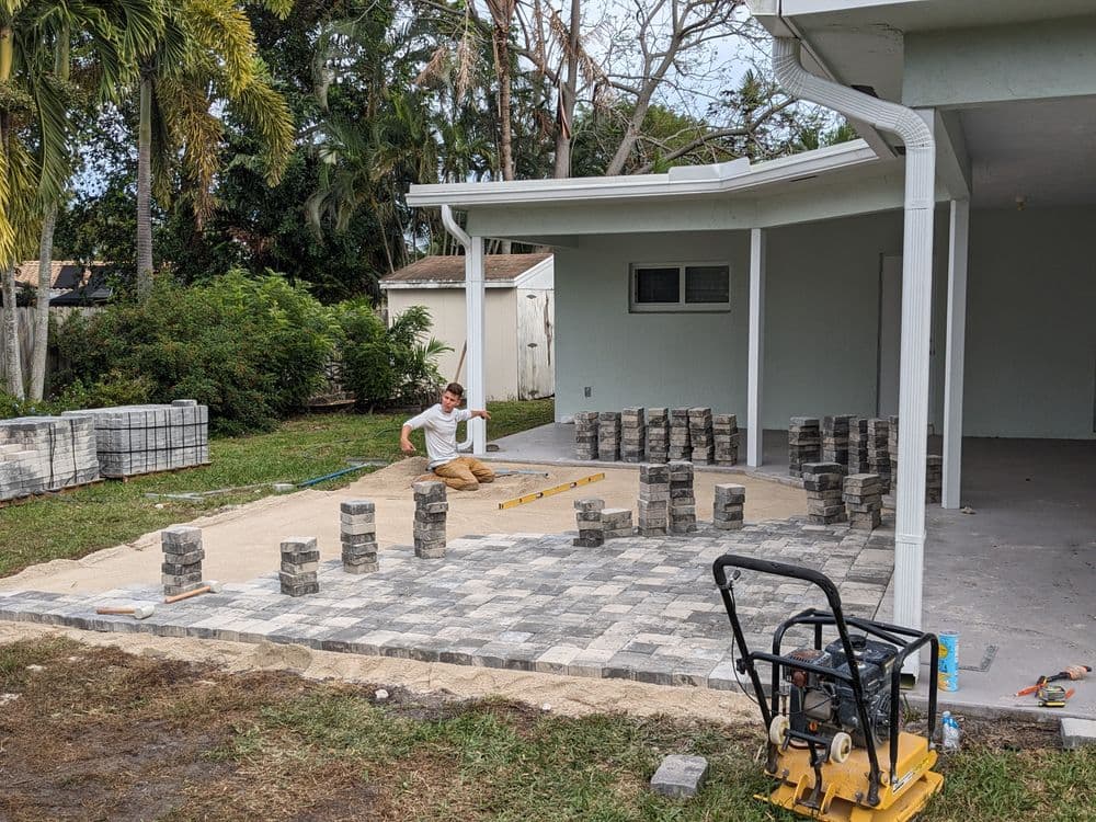 Construction worker laying paver stones in backyard with tools and materials nearby.