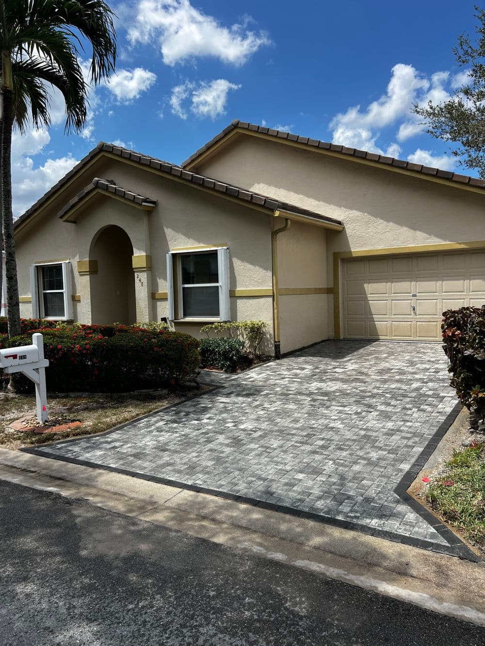 Single-story home with paver driveway, mailbox, and landscape near palm trees under blue sky.