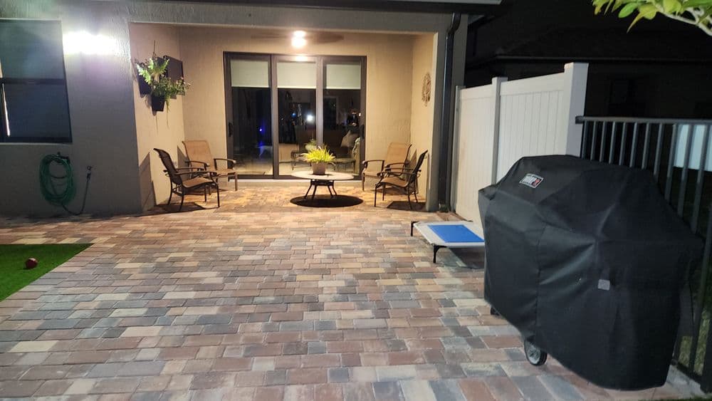 Patio with paver stones, chairs, table, and a covered grill in a well-lit outdoor space.