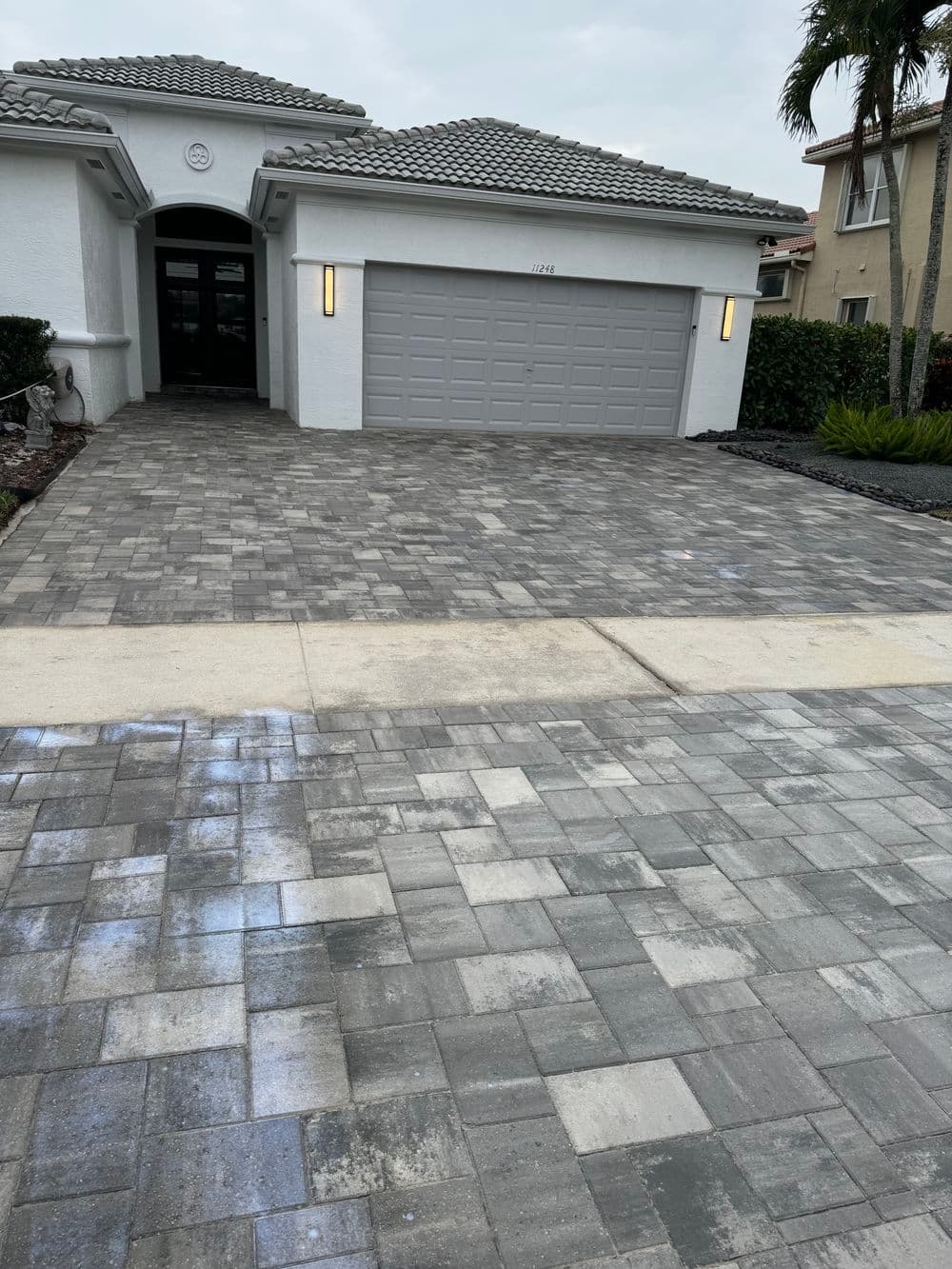 Modern house with a gray paver driveway and garage door, surrounded by landscaped greenery.