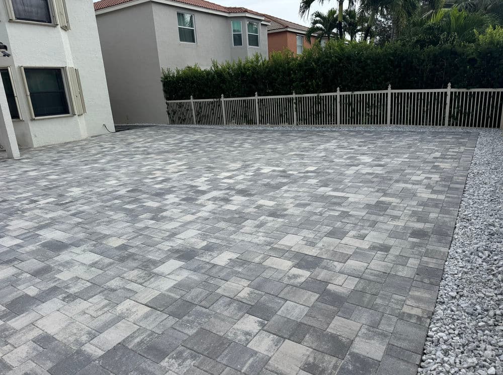 Paved outdoor patio area with gray bricks, surrounded by greenery and a fence.