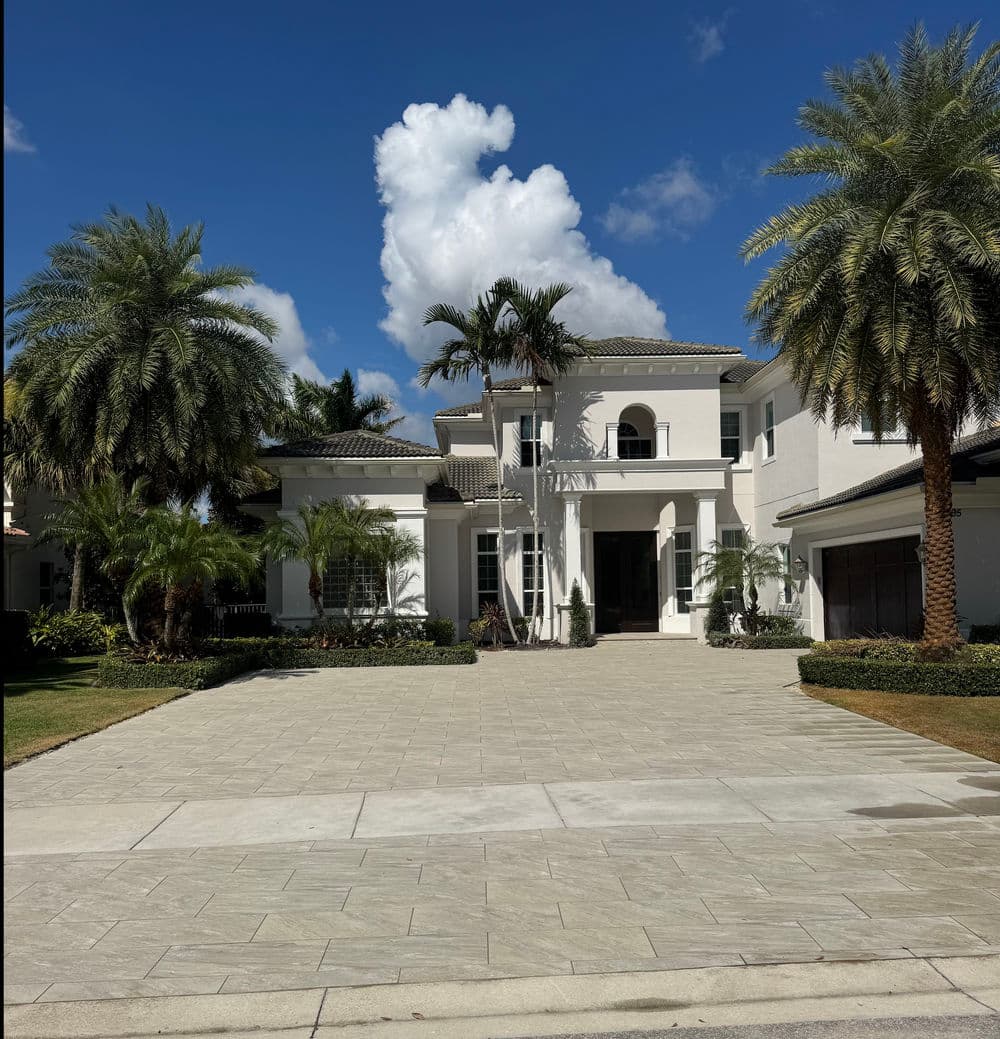 Modern luxury home with palm trees and blue sky, featuring a spacious driveway and white facade.